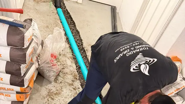 Technician shovelling gravel around new green PVC drain pipe and fittings in an open basement floor trench, stone mix bags stacked beside him